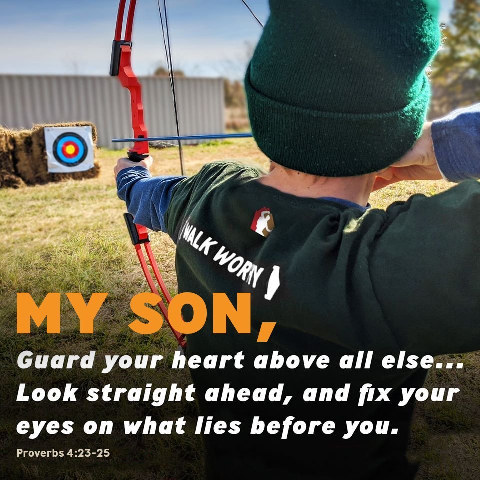 Boy aiming a bow at an outdoor archery target during troop activity.